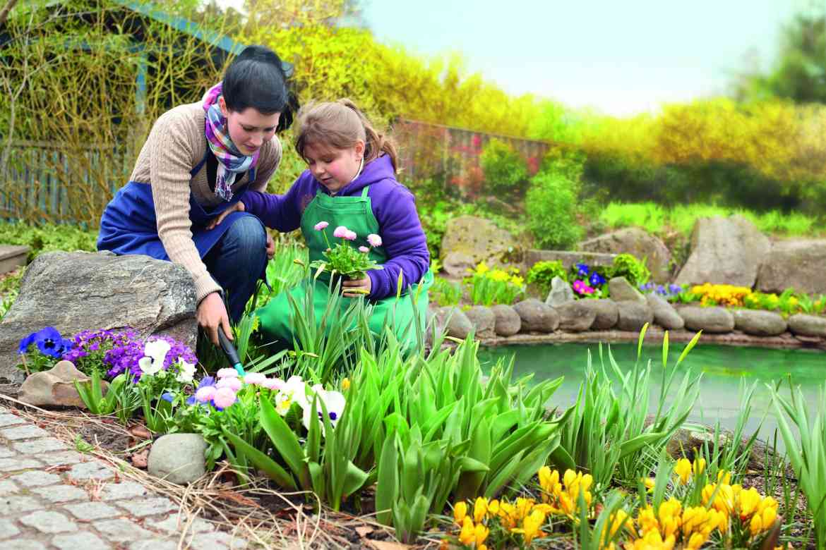 Eine junge ASB-Freiwillige pflanzt mit einem Mädchen Blumen an einem Gartenteich.