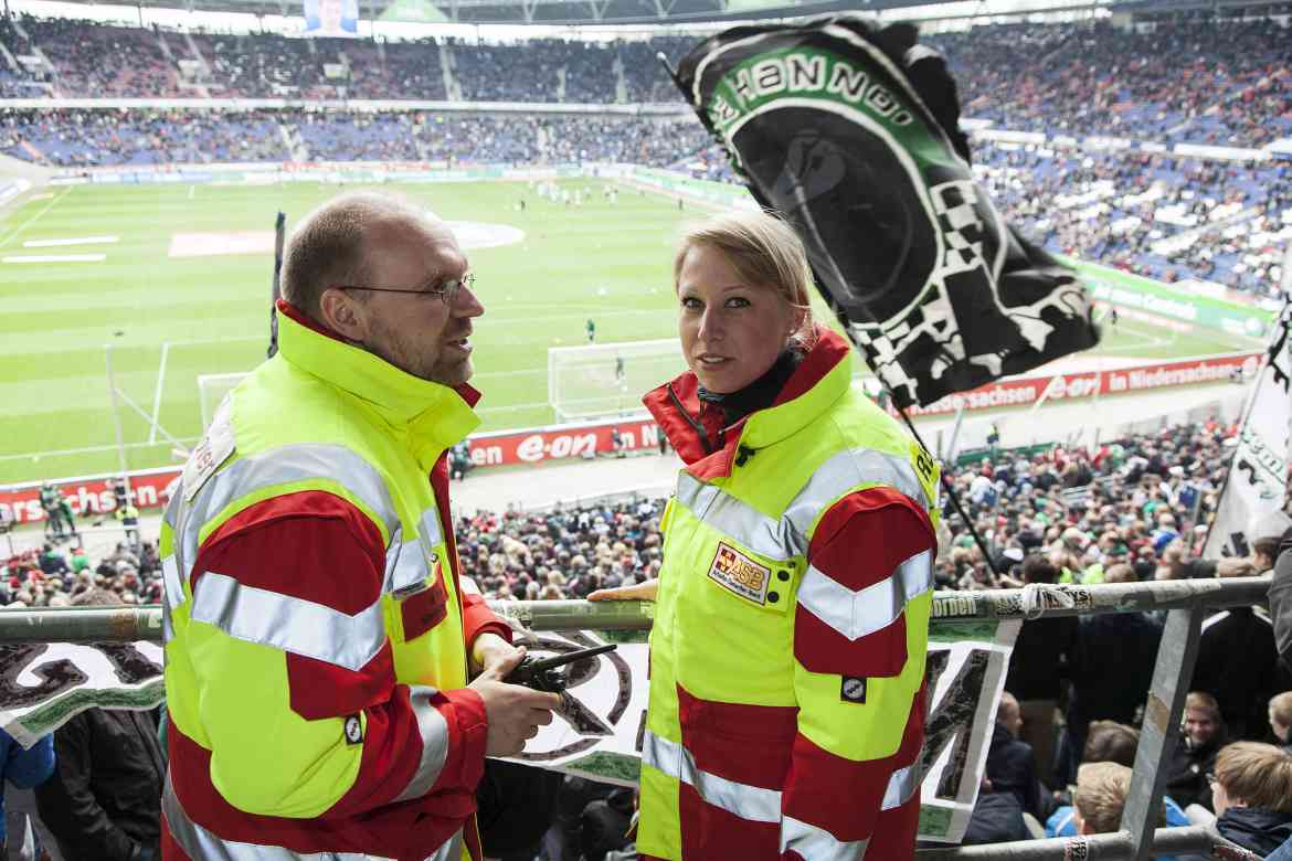 Ein Sanitätshelfer und eine Sanitätshelferin stehen in einem vollbesetzten Fussballstadion.