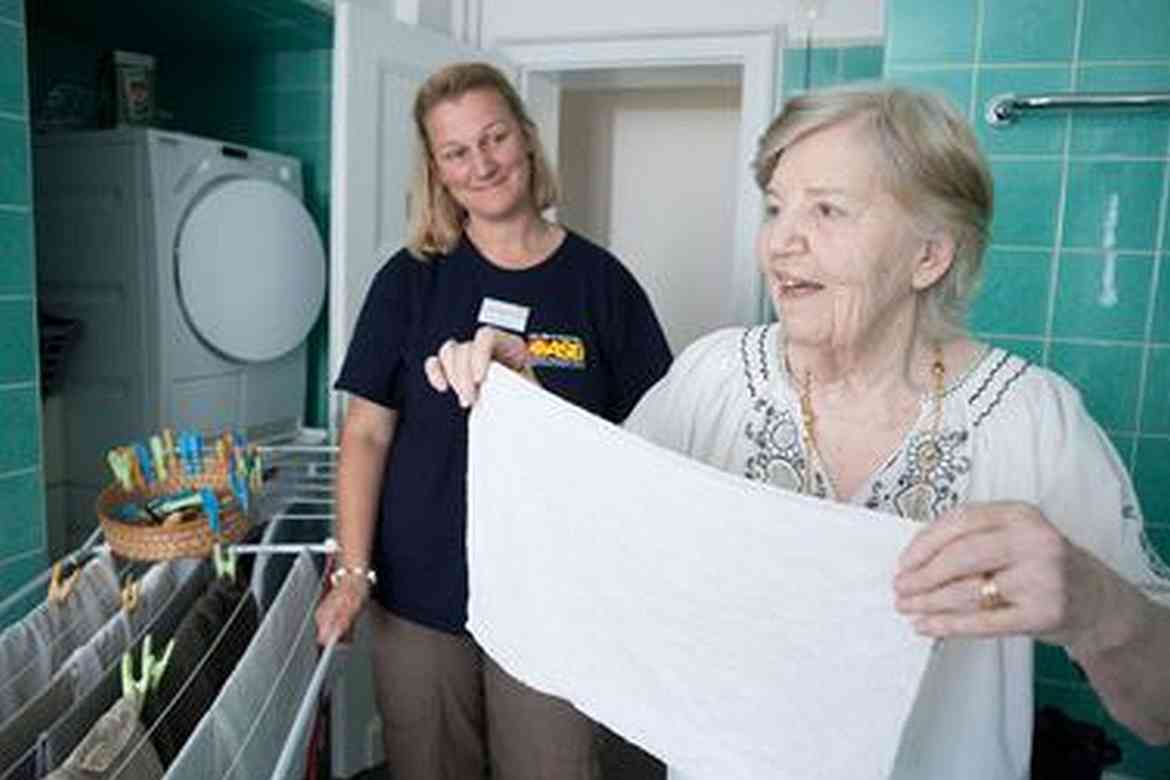 Eine ASB-Pflegerin beugt sich lächelnd zu einer Seniorin,die an ihrem Esstisch Kaffee trinkt.
