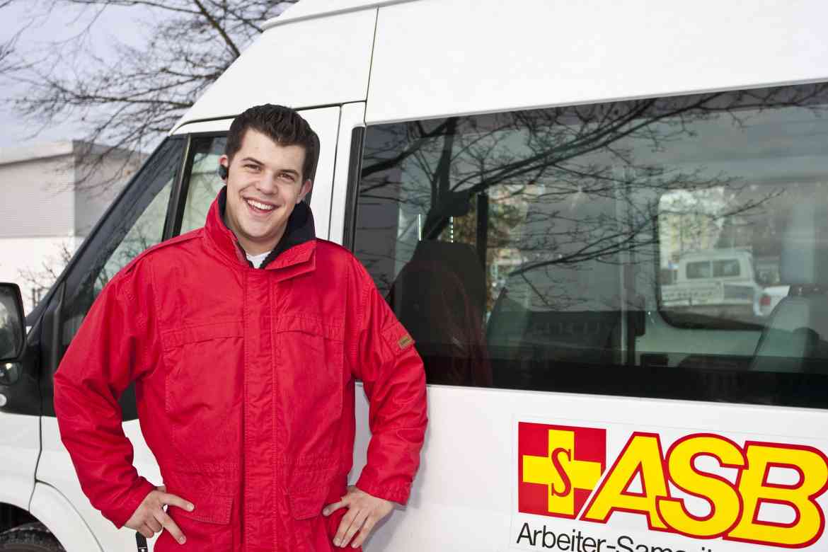 ASB Mitarbeiter steht lächelnd vor seinem Dienstfahrzeug.