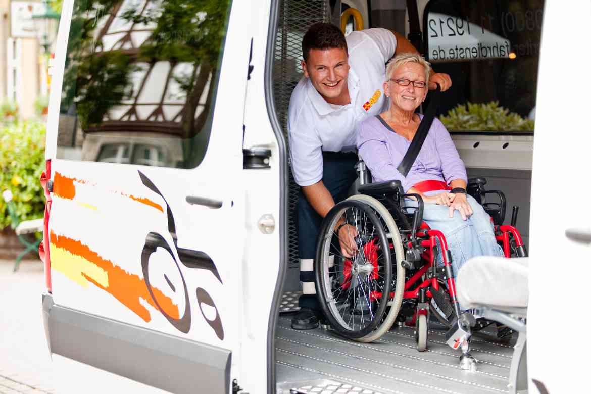 Ein Fahrdienstmitarbeiter hilft Frau im Rollstuhl aus dem Fahrzeug.