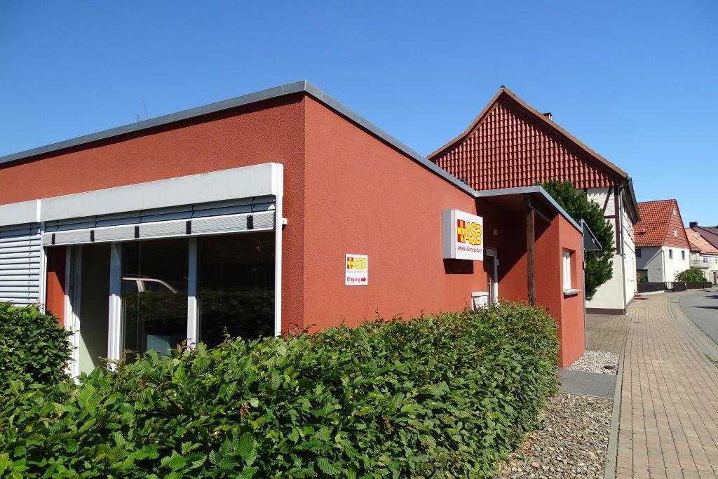 Aussenansicht des ASB in Wulften. Ein rotes Gebäude mit großen Fenstern, davor eine grüne Hecke, im Hintergrund ein blauer Himmel